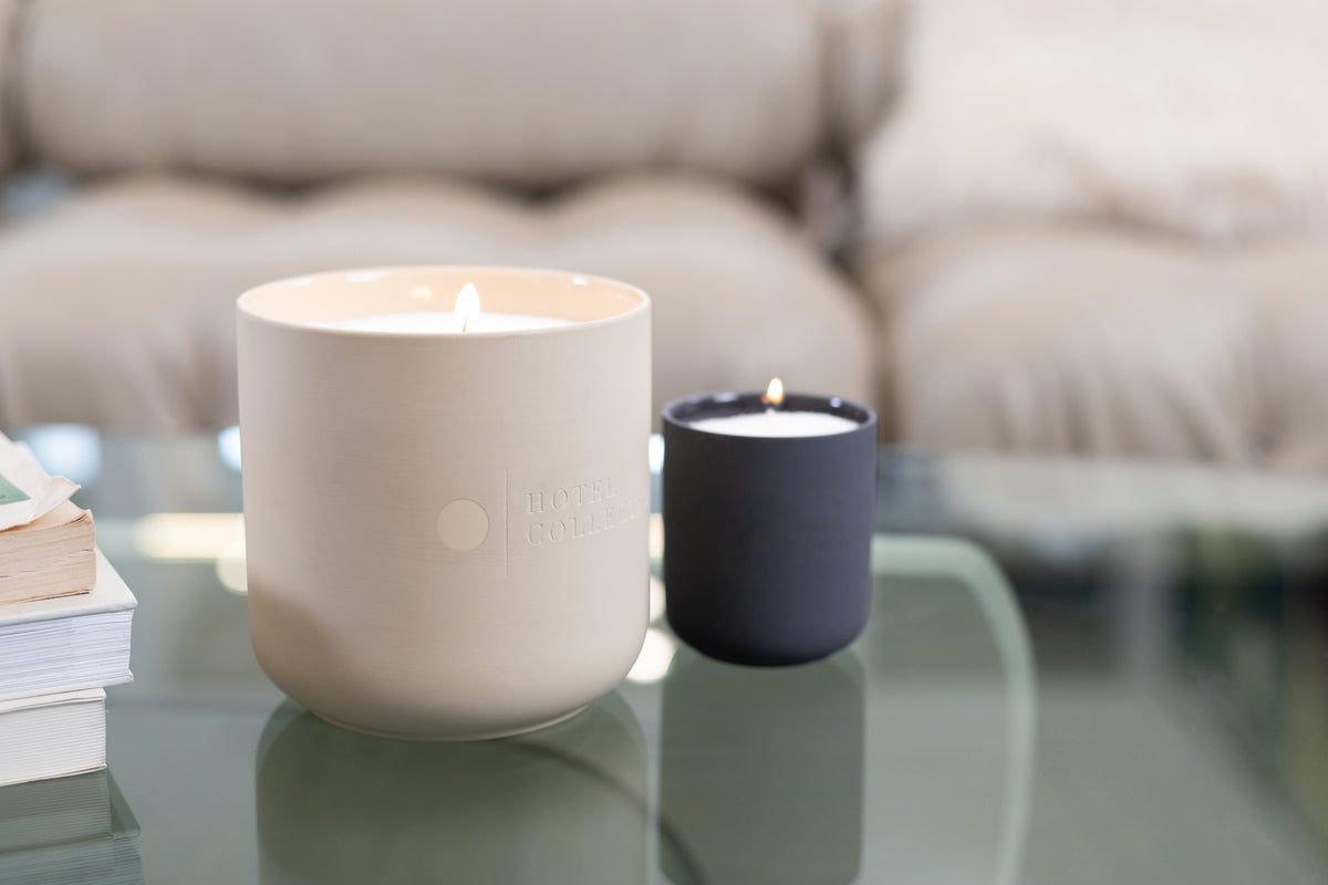 Two lit sand wax candles from Hotel Collection are displayed on a glass coffee table in a cozy living room. The larger candle is in a matte bone vessel, and the smaller one is in a matte charcoal vessel.